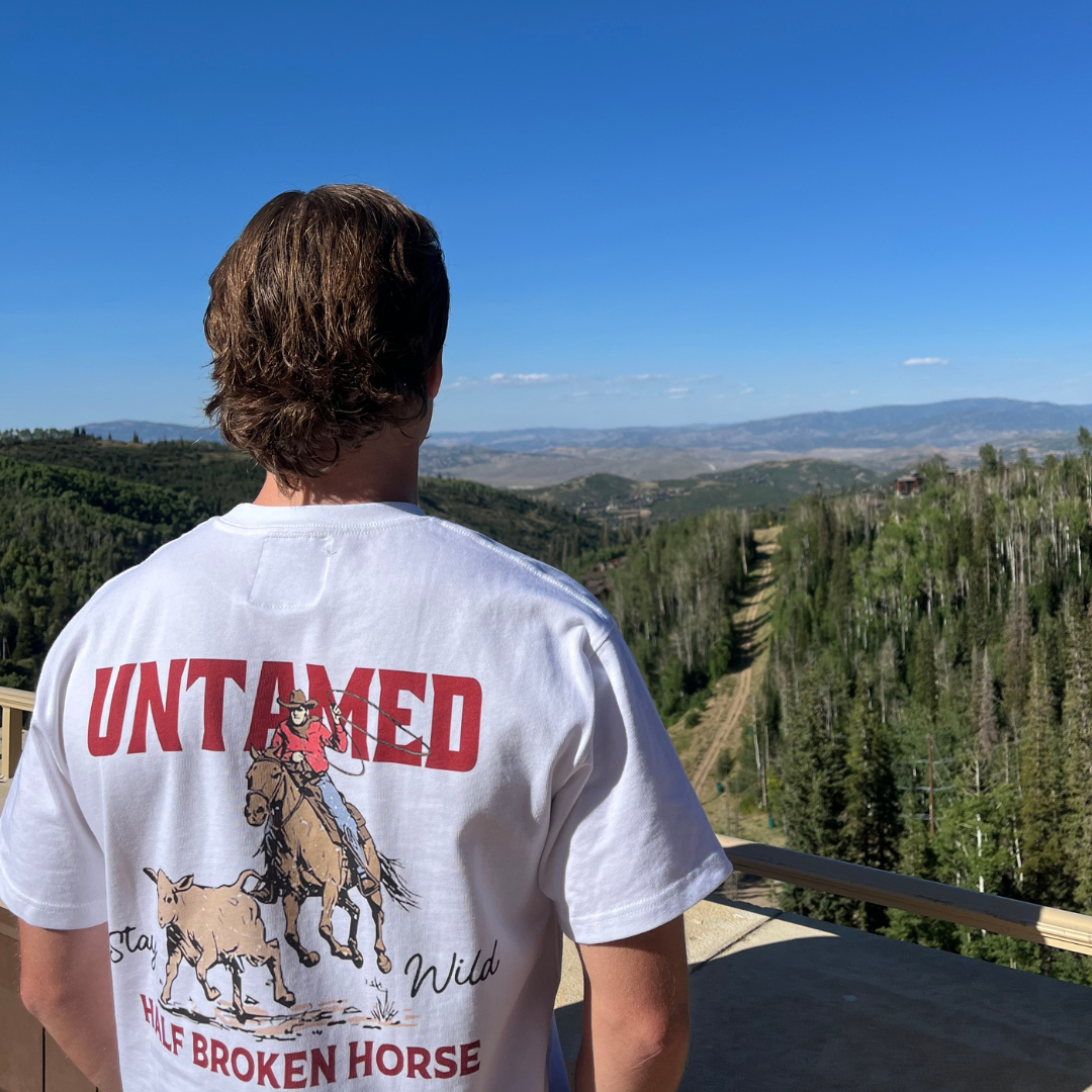 Person wearing a white t-shirt with a graphic and text, standing on a wooden deck with a scenic view of trees and mountains.
