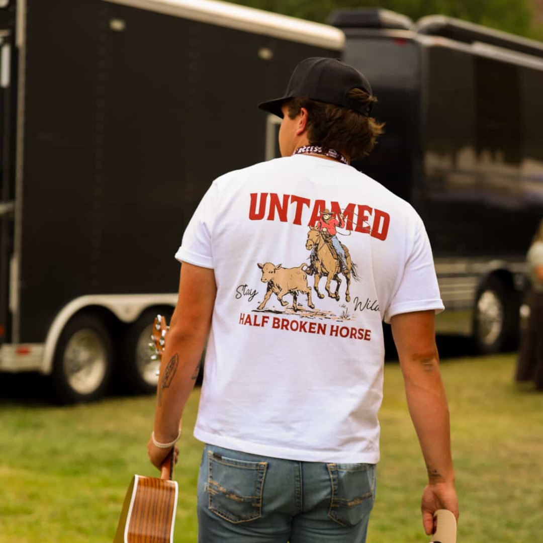 Person wearing a white t-shirt with 'Untamed' and 'Half Broken Horse' text, standing outdoors with trailers in the background.